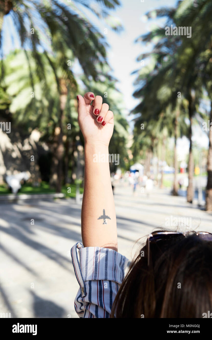 Tatouage d'un avion dans l'avant-bras d'une fille millénaire à l'extérieur dans la rue entourée de palmiers en été. Banque D'Images