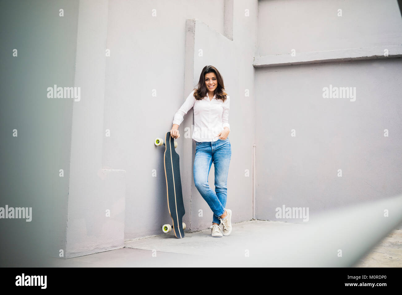 Smiling young woman with skateboard leaning against wall sur terrasse Banque D'Images