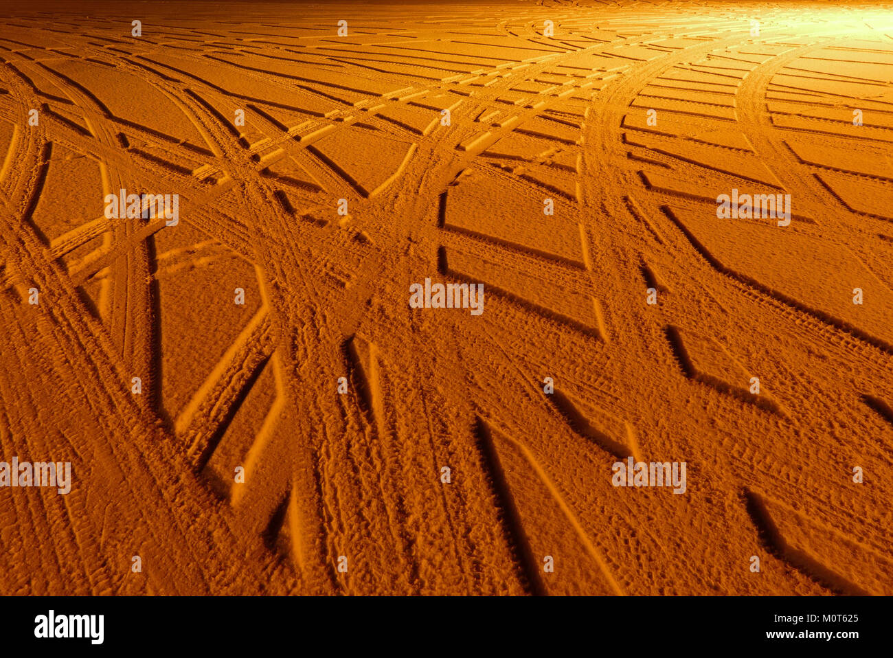 L'image capture les traces de pneus de voiture dans la neige fraîche, éclairées par la lueur chaude des lampadaires au sodium, soulignant le contraste entre la texture de la neige et l'environnement d'éclairage urbain. Banque D'Images