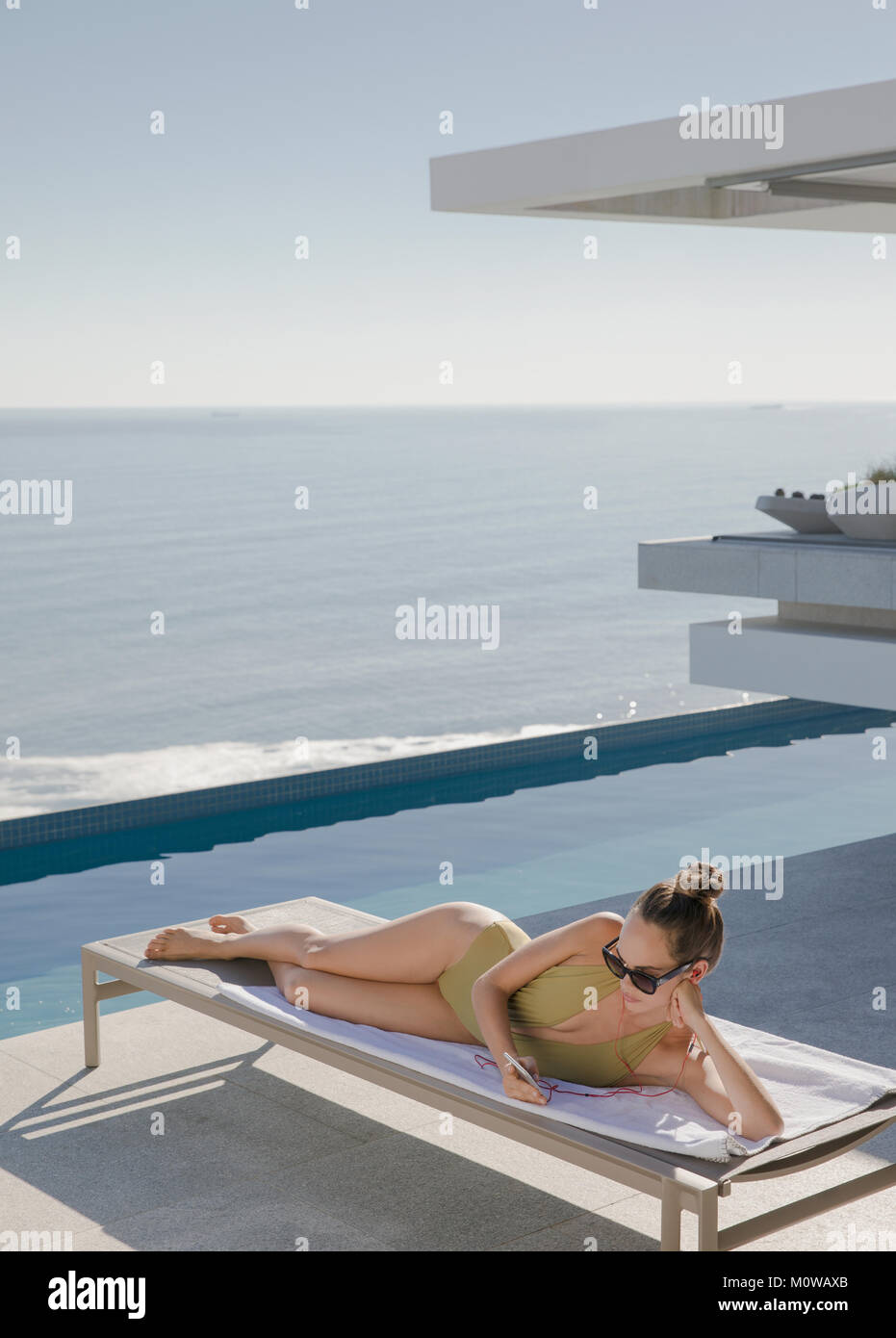 Femme de soleil, texting with smart phone on lounge chair on sunny patio avec vue sur l'océan de luxe Banque D'Images