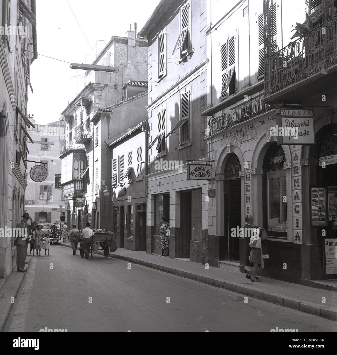 Années 1950, historique vue vers le bas d'une rue près de Governor's Street, Gibraltar, montrant un marchand et le Medical Hall, avec produits Boots the Chemist, population locale et panier. Architecture Espagnole avec quelques habitants de la signalisation et des noms. Banque D'Images