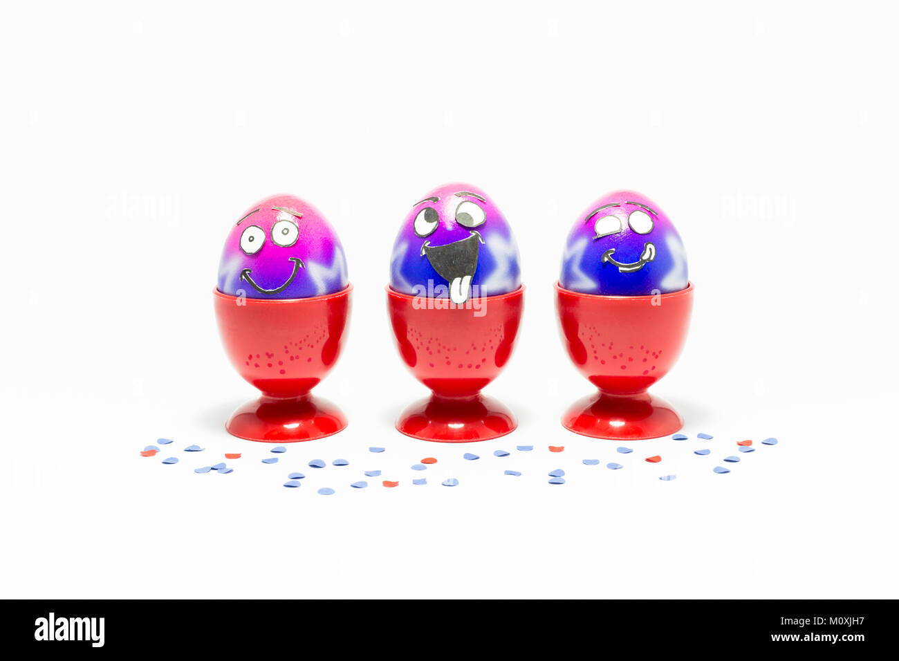 Groupe d'oeufs de Pâques peints colorés avec drôle cartoon style visages dans des tasses et des oeufs en plastique rouge confetti colorés sur fond blanc Banque D'Images