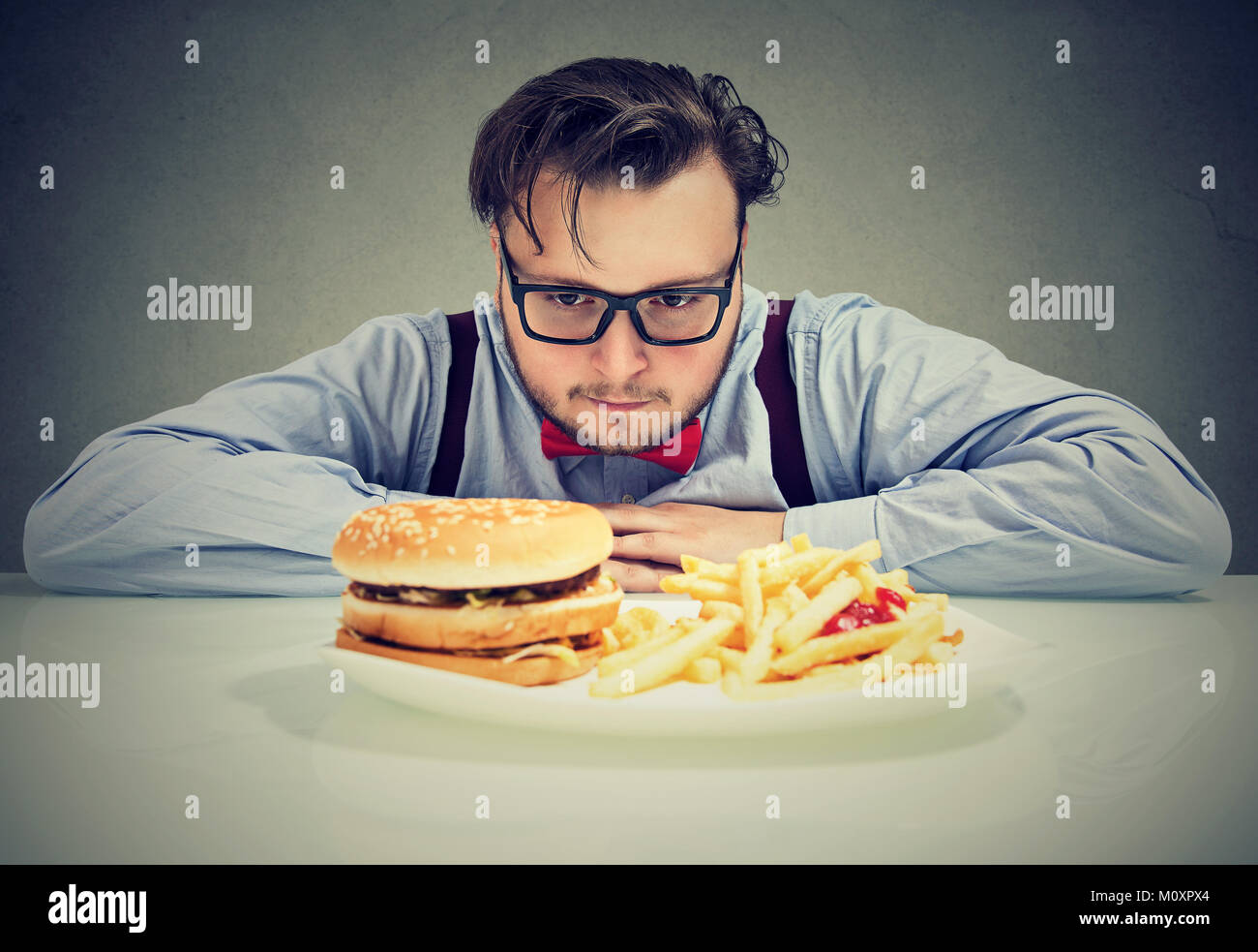 Jeune homme à lunettes chunky envie cheeseburger avec frites à souligné tout en étant sur l'alimentation. Banque D'Images
