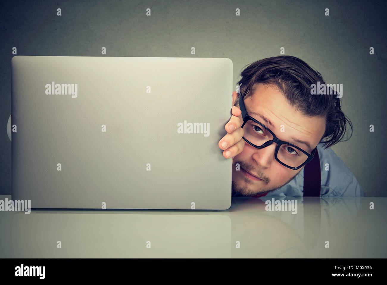 Jeune homme fatigué lunettes en essayant de se cacher derrière un ordinateur portable à la peur de la concurrence. Banque D'Images