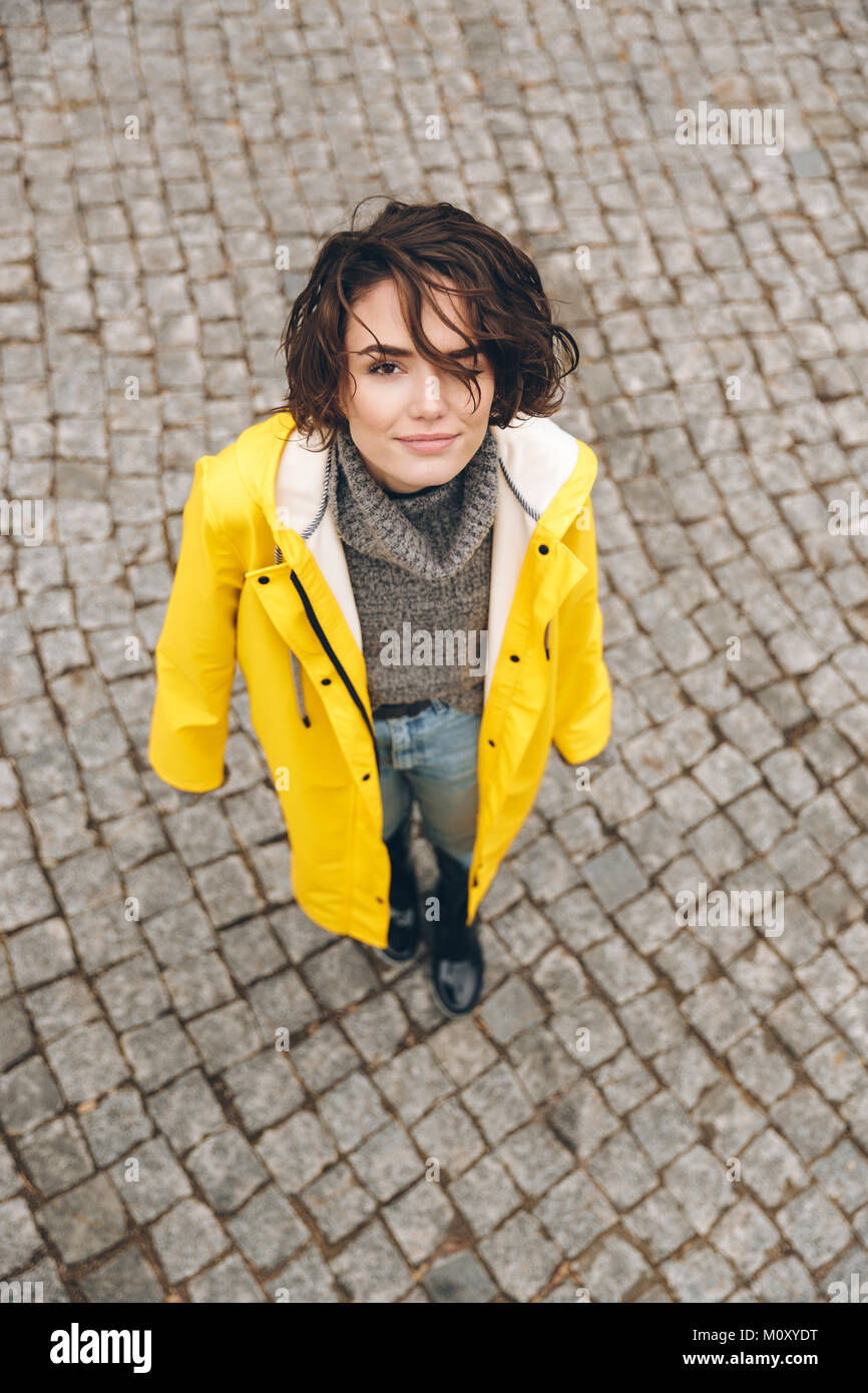 Portrait de mignon femelle jaune en manteau élégant à la recherche jusqu'à la caméra, en se tenant sur le pavement au cours de sa promenade Banque D'Images