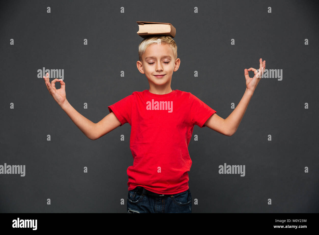 Photo de petit garçon enfant isolé sur fond gris méditer avec livre sur la tête. Banque D'Images