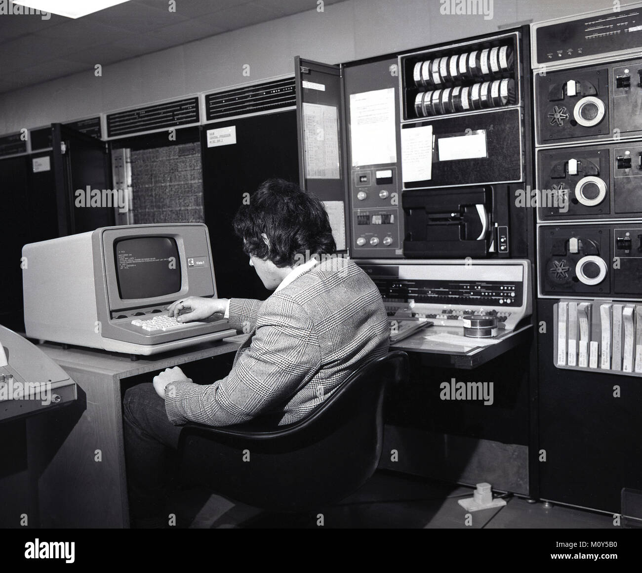 70, tableau historique d'un homme dans une salle de traitement des données avec des banques de matériel électronique à côté de lui, assis à une petite station de travail à l'aide d'un ordinateur avec écran, England, UK. Banque D'Images