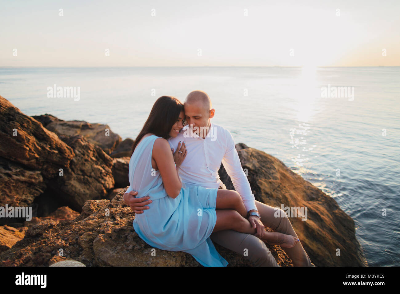 Caucasian couple hugging on rocks sur la plage au coucher du soleil Banque D'Images