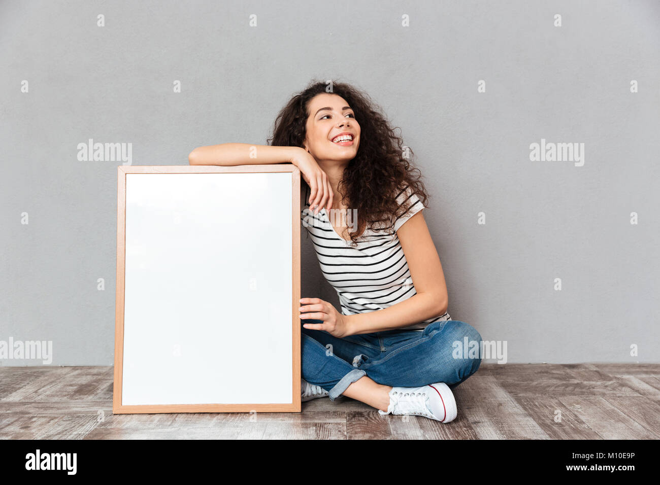 Femme de race blanche avec de beaux cheveux qui posent avec les jambes croisées la démonstration de grande grande peinture ou portrait, isolé sur mur gris copy space Banque D'Images