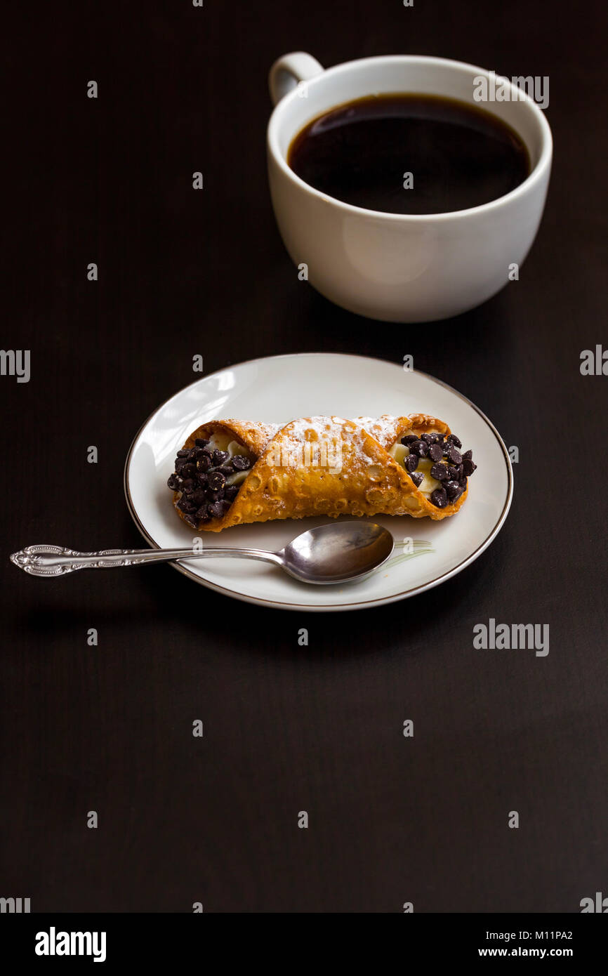 Cannoli italiens avec des copeaux de chocolat sur plaque blanche et café tasse sur fond noir Banque D'Images