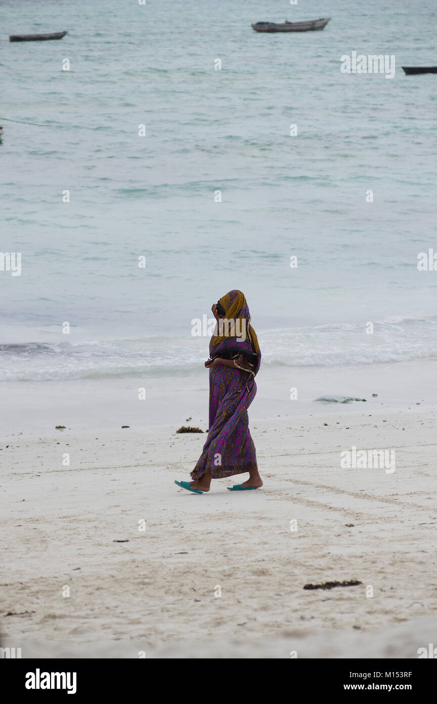 Femme musulmane marche sur la plage de Nungwi, Zanzibar, couvertes de la tête aux pieds et portant un foulard sur une journée ensoleillée à la plage, les dhows derrière elle Banque D'Images