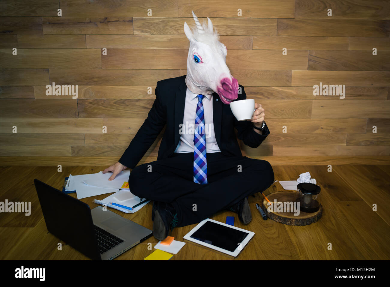 Licorne en costume et cravate sourit et boit du café. Jeune homme drôle de  masque en caoutchouc est assis sur le sol contre un mur et travaille au  bureau à domicile Photo
