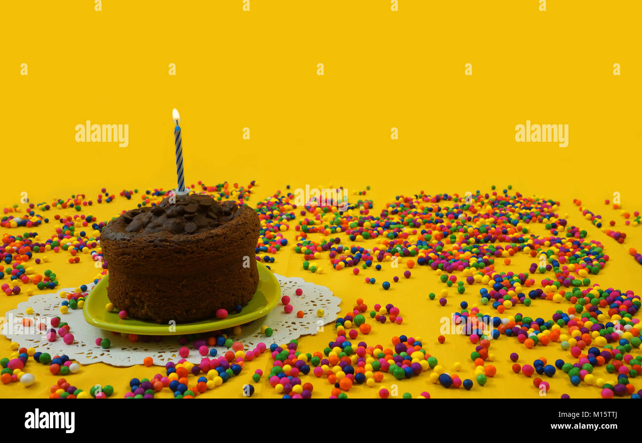 Gâteau au chocolat avec 1 bleu et blanc bougie allumée, sur une petite plaque verte, entourée par candy balls éparpillées sur un fond jaune Banque D'Images