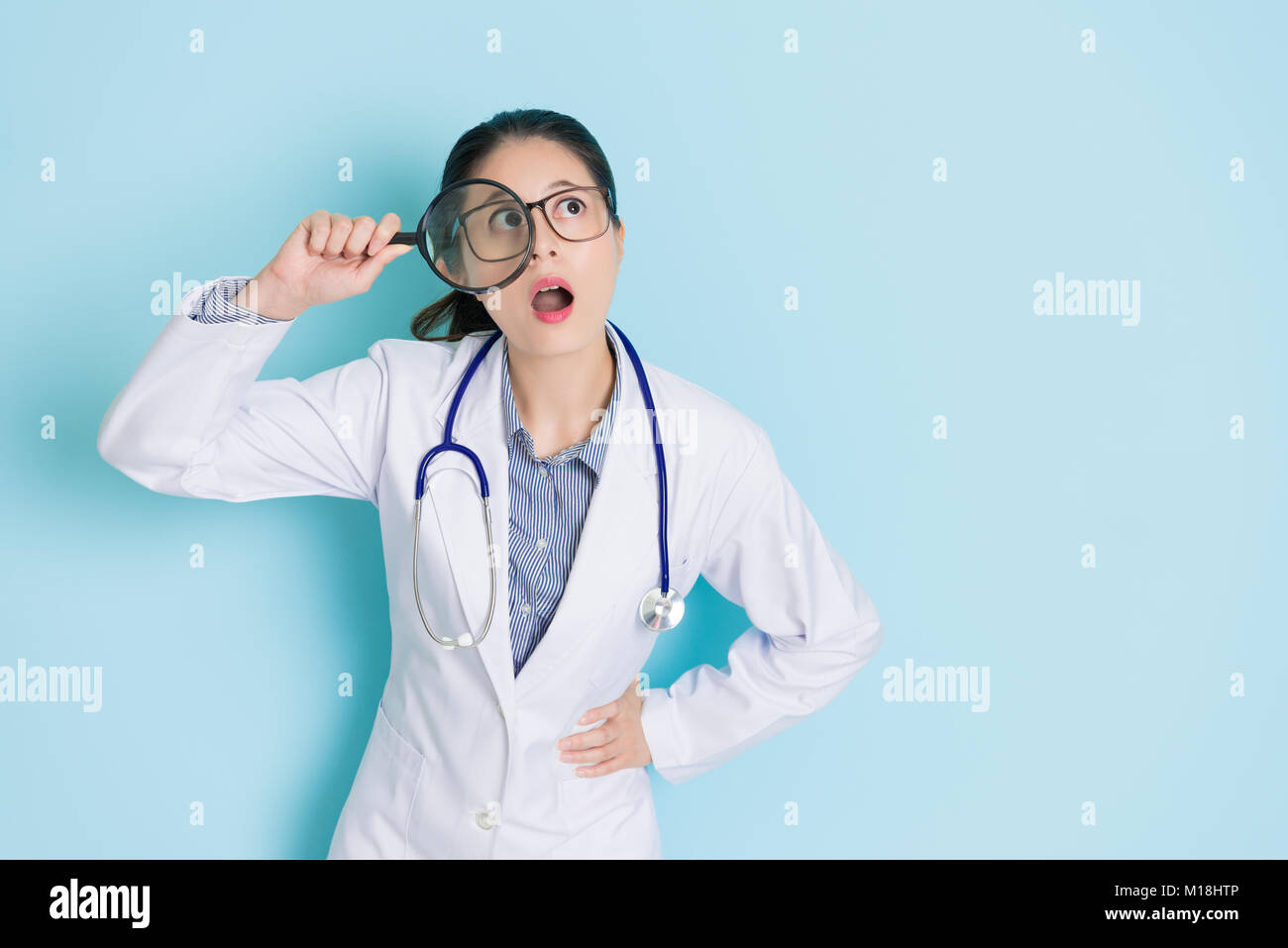 Young female doctor standing in fond bleu à l'aide de loupe à l'air de penser à une solution de soins et avoir une bonne idée de se sentir choqué. Banque D'Images Young female doctor standing in fond bleu à l'aide de loupe à l'air de penser à une solution de soins et avoir une bonne idée de se sentir choqué. Banque D'Images