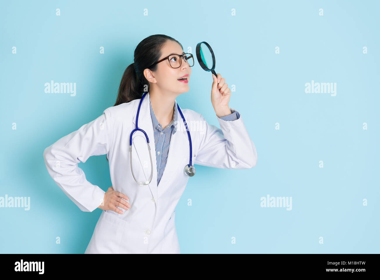 Beauté smiling girl médecin exerçant son stéthoscope debout dans le fond bleu et l'utilisation de l'outil loupe à la recherche ci-dessus à l'information médicale. Banque D'Images Beauté smiling girl médecin exerçant son stéthoscope debout dans le fond bleu et l'utilisation de l'outil loupe à la recherche ci-dessus à l'information médicale. Banque D'Images