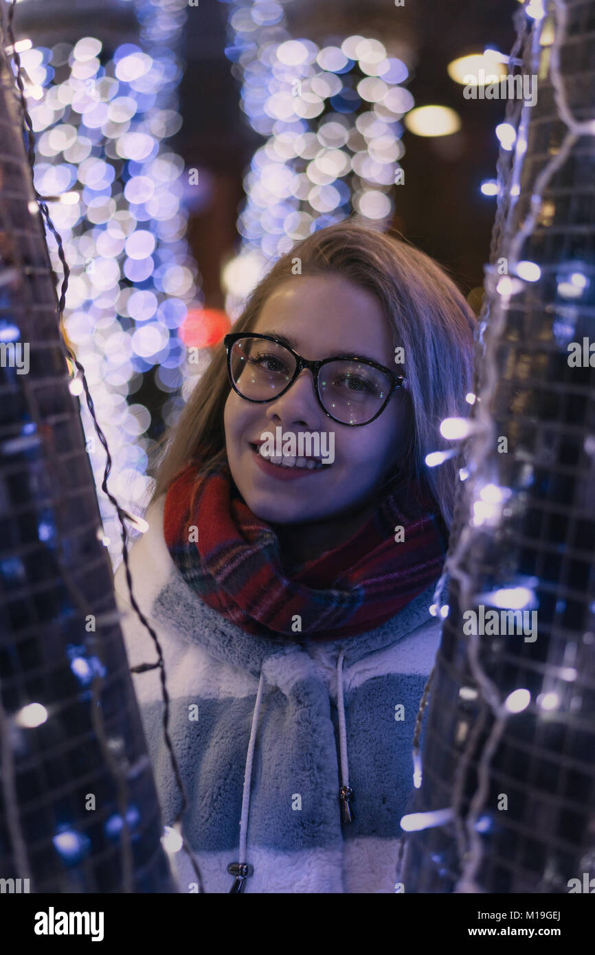 Jeune fille attirante dans des verres à pied à la ville est juste. Elle sourit. Portrait de nuit entre les guirlandes. Banque D'Images