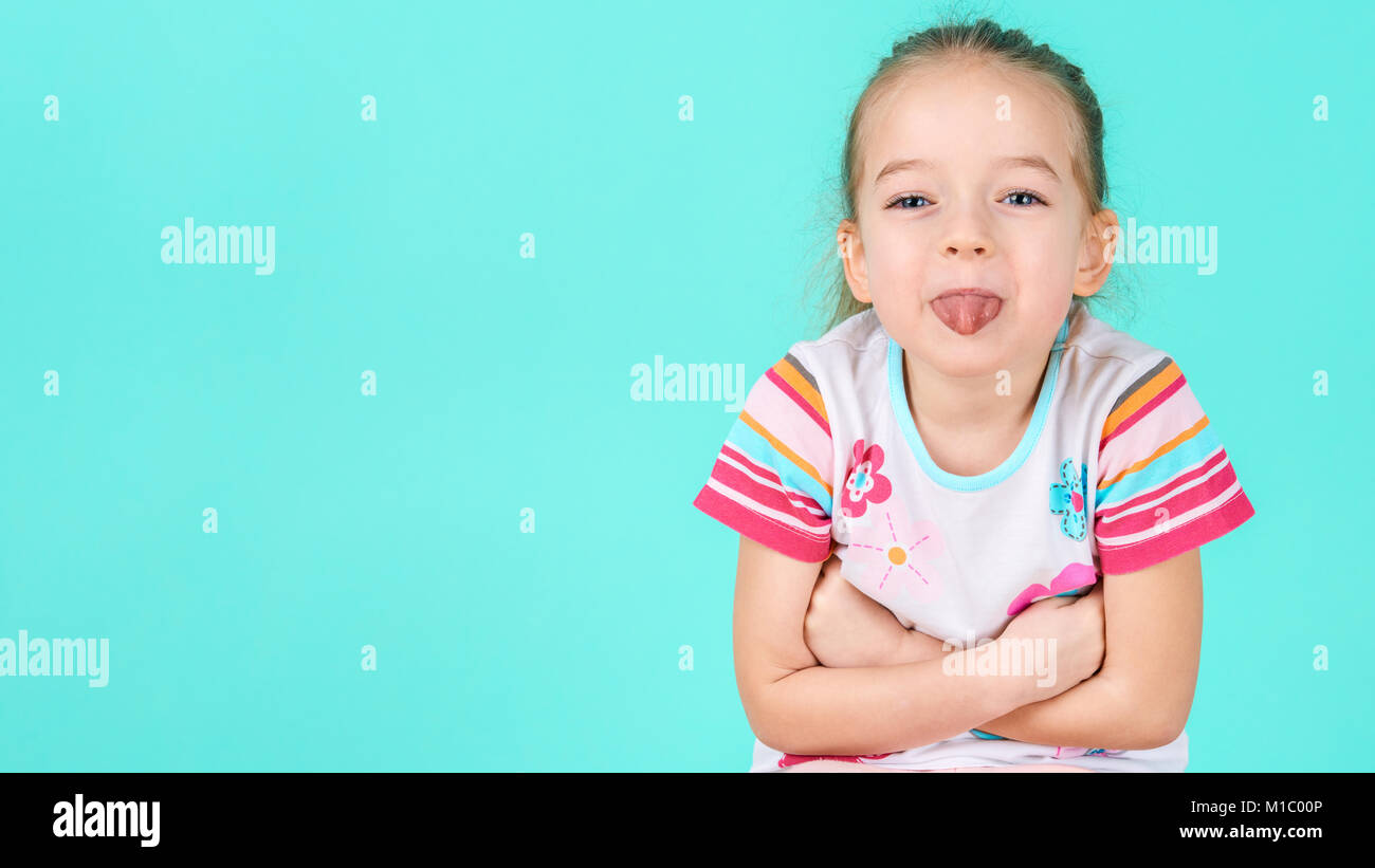 Adorable petite fille aux bras croisés coller sa langue dehors. Cheeky bambin taille portrait sur fond bleu candy couleur pastel. Banque D'Images