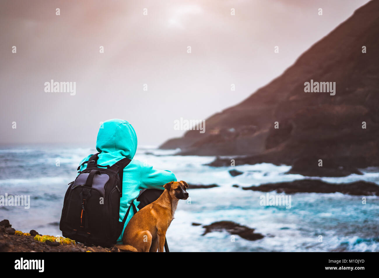 Homme avec un chien en face de paysage littoral rural avec des montagnes et des vagues venant et rayons de soleil à travers les nuages. L'île de Santo Antao, Cap Vert Banque D'Images