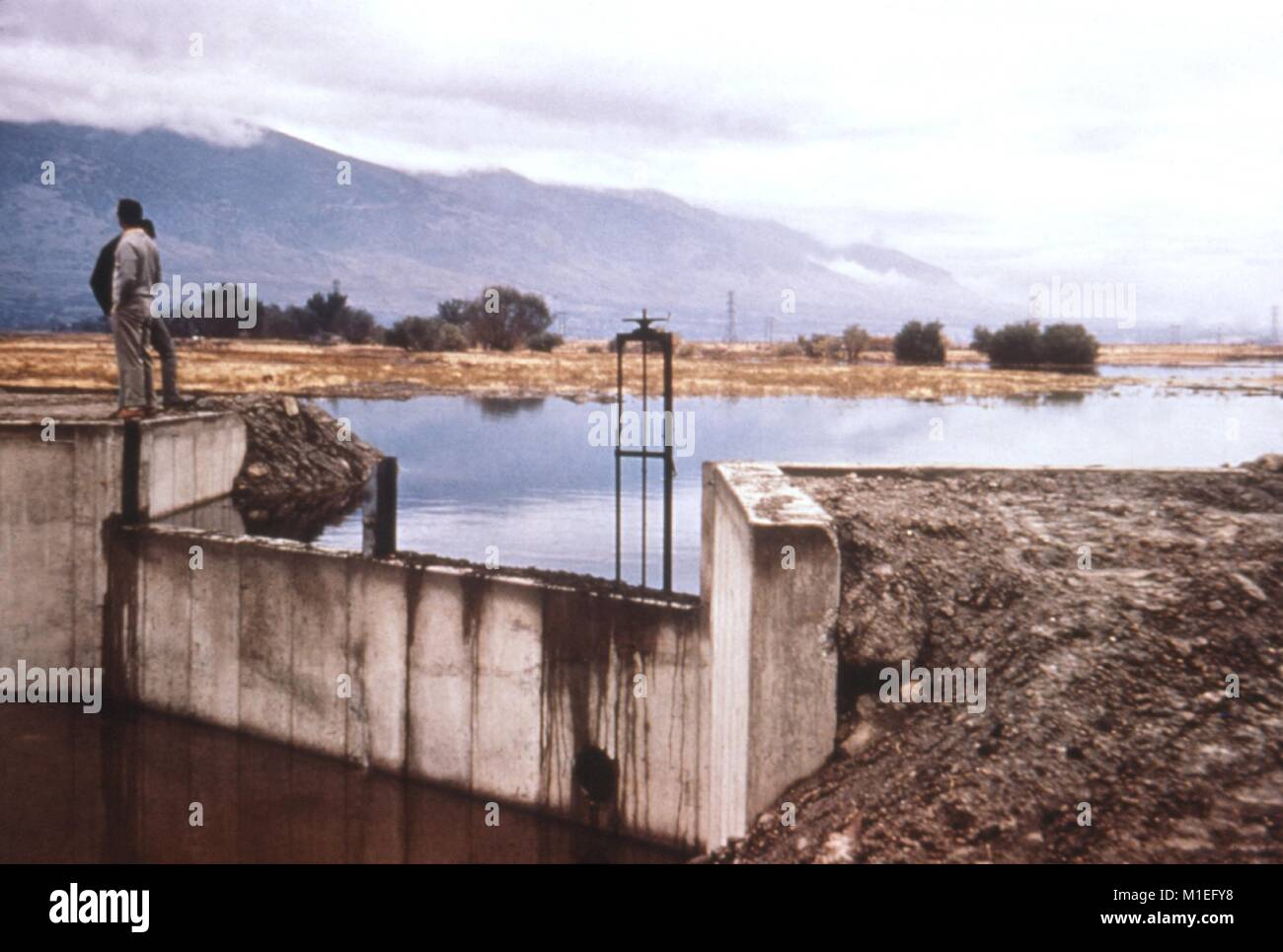 Photographie montrant une vue avec une vanne en béton à un réservoir d'eau, avec deux personnes debout à côté d'elle et un paysage montagneux en arrière-plan, une position de l'enquête CDC pour minimiser les risques de maladies transmises par vecteur autour de l'évolution des ressources en eau, 1976. Image courtoisie CDC. () Banque D'Images Photographie montrant une vue avec une vanne en béton à un réservoir d'eau, avec deux personnes debout à côté d'elle et un paysage montagneux en arrière-plan, une position de l'enquête CDC pour minimiser les risques de maladies transmises par vecteur autour de l'évolution des ressources en eau, 1976. Image courtoisie CDC. () Banque D'Images