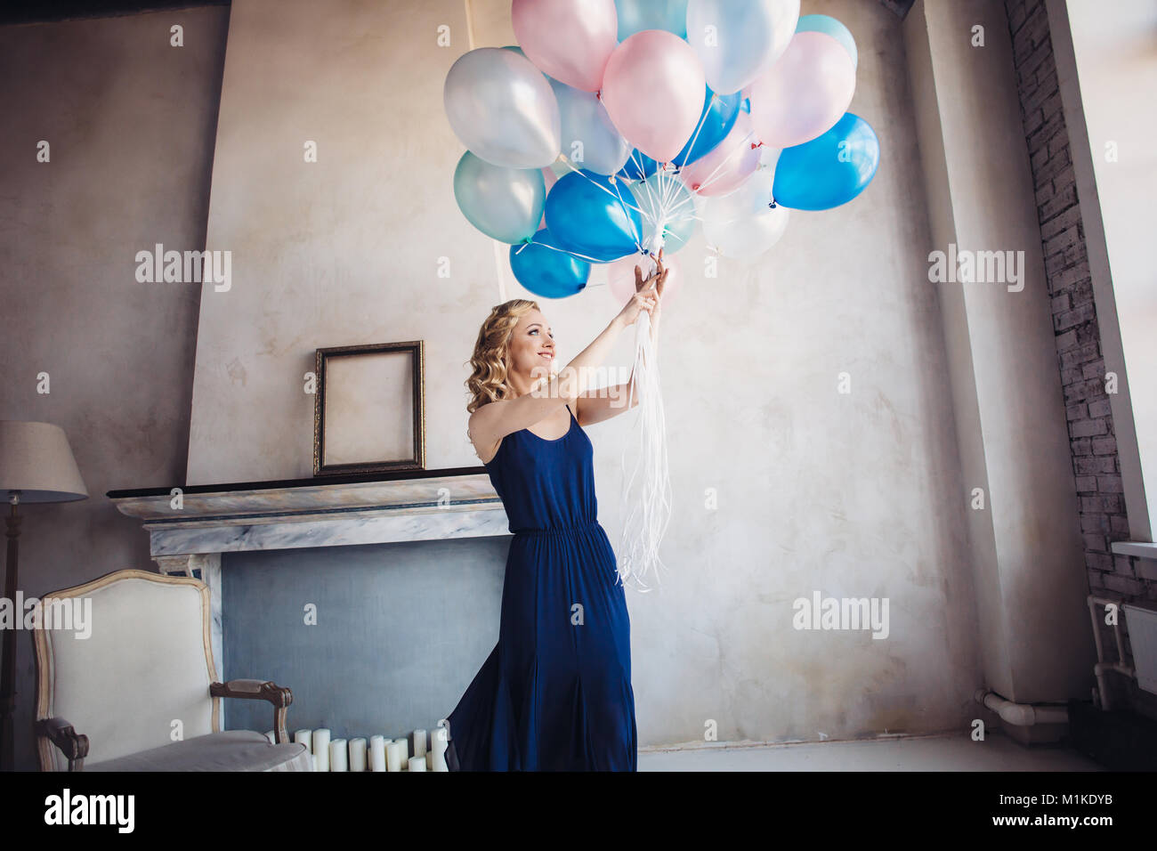 Femme blonde en robe bleu avec bleu et rose baloons regarder baloons et sourires, sur fond intérieur minimaliste Banque D'Images