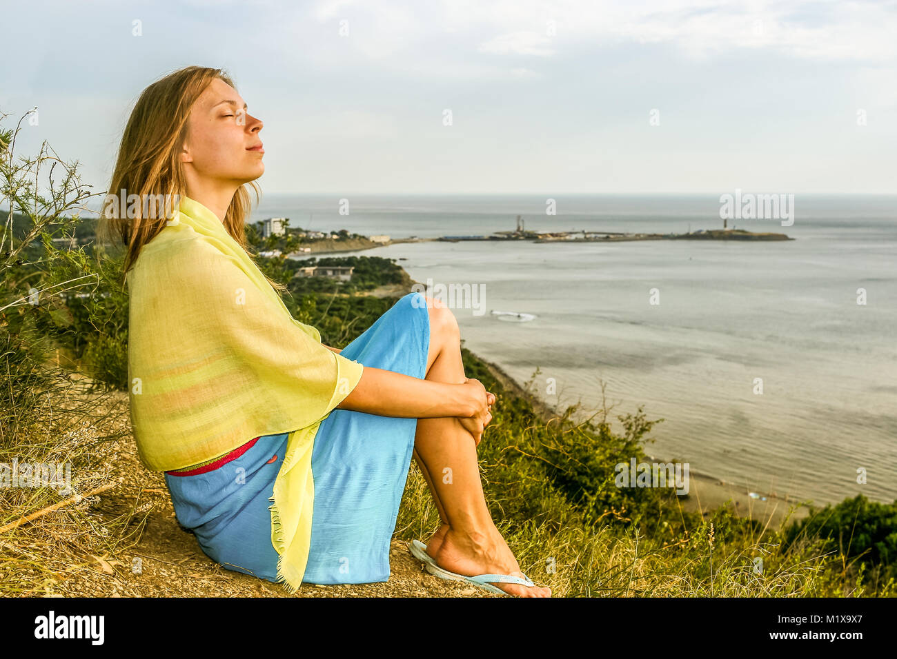 Jeune fille aux yeux clos est assis sur une haute côte au-dessus de la mer et jouit le soleil du soir. Banque D'Images