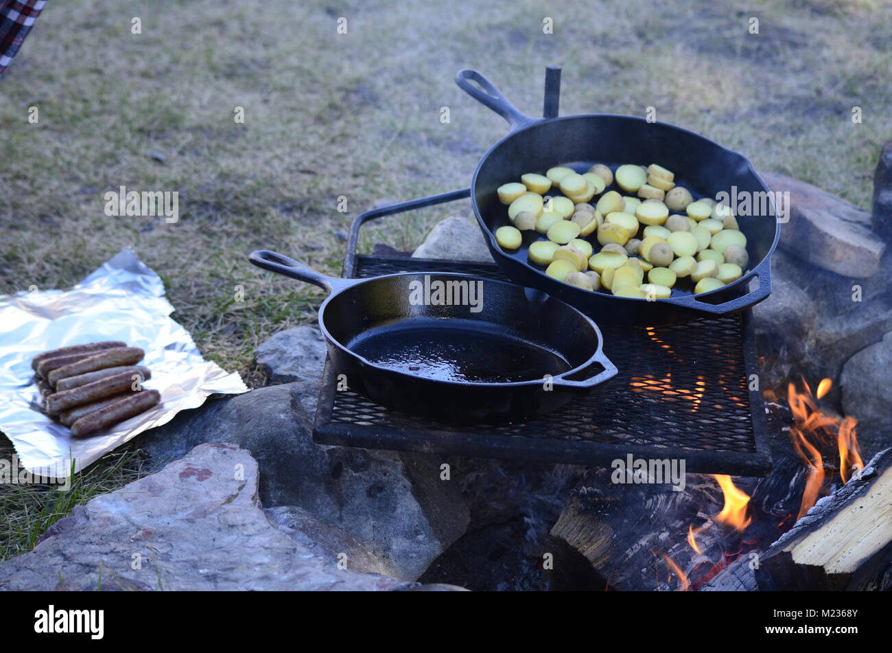 Deux poêles en fonte, s'asseoir sur une grille au-dessus d'un feu chaud. La cuisson d'un petit-déjeuner tôt le matin avec petit déjeuner saucisses et pommes de terre en tranches Banque D'Images