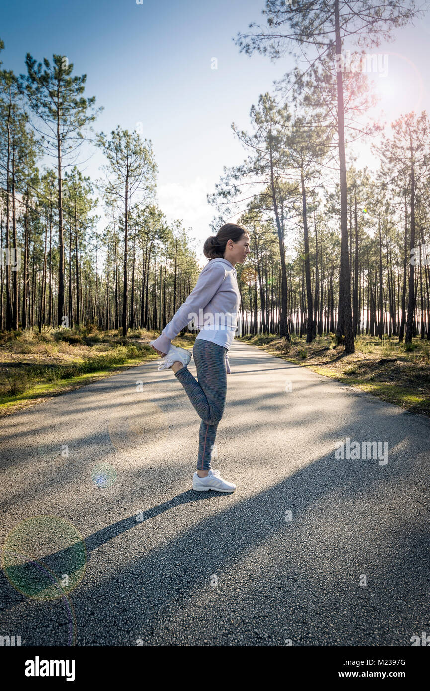 Femme jogger faisant une extension des jambes jusqu'au milieu d'une route de campagne qui traverse une forêt. Banque D'Images