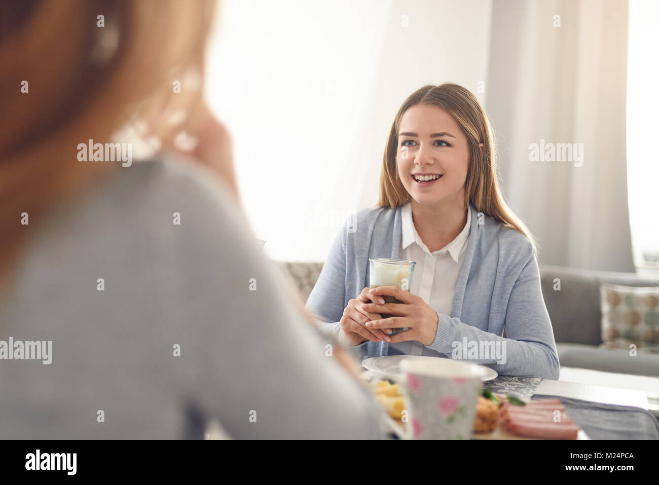 Happy smiling adolescent ayant le petit déjeuner à la maison assis à la table avec une boisson et de la transmission à la personne en face qui est hors de l'écran Banque D'Images