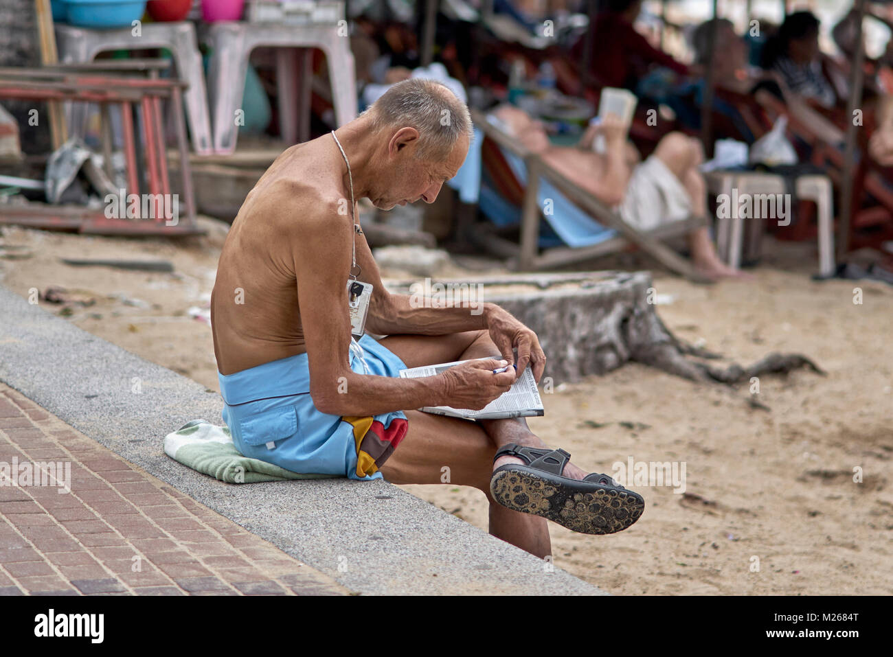 Homme sans chirte à la plage faisant un puzzle de mots croisés de journal. Thaïlande, Asie du Sud-est Banque D'Images