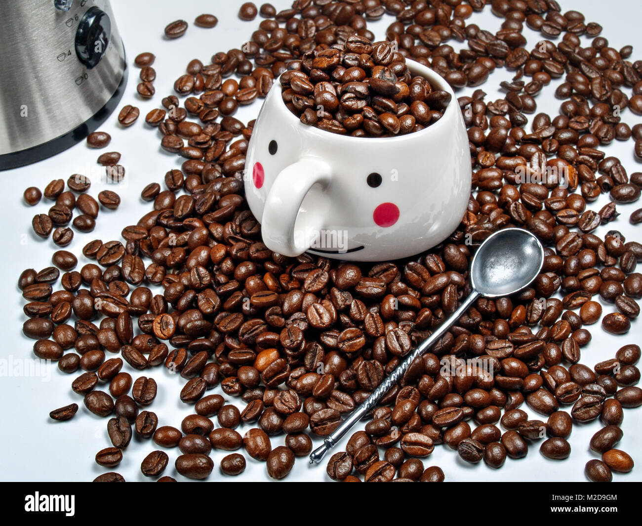 Tasse de café blanc rempli de grains de café est sur un tas de fèves de café torréfié à côté d'une cuillère en métal se trouve et il y a un moulin à café électrique Banque D'Images