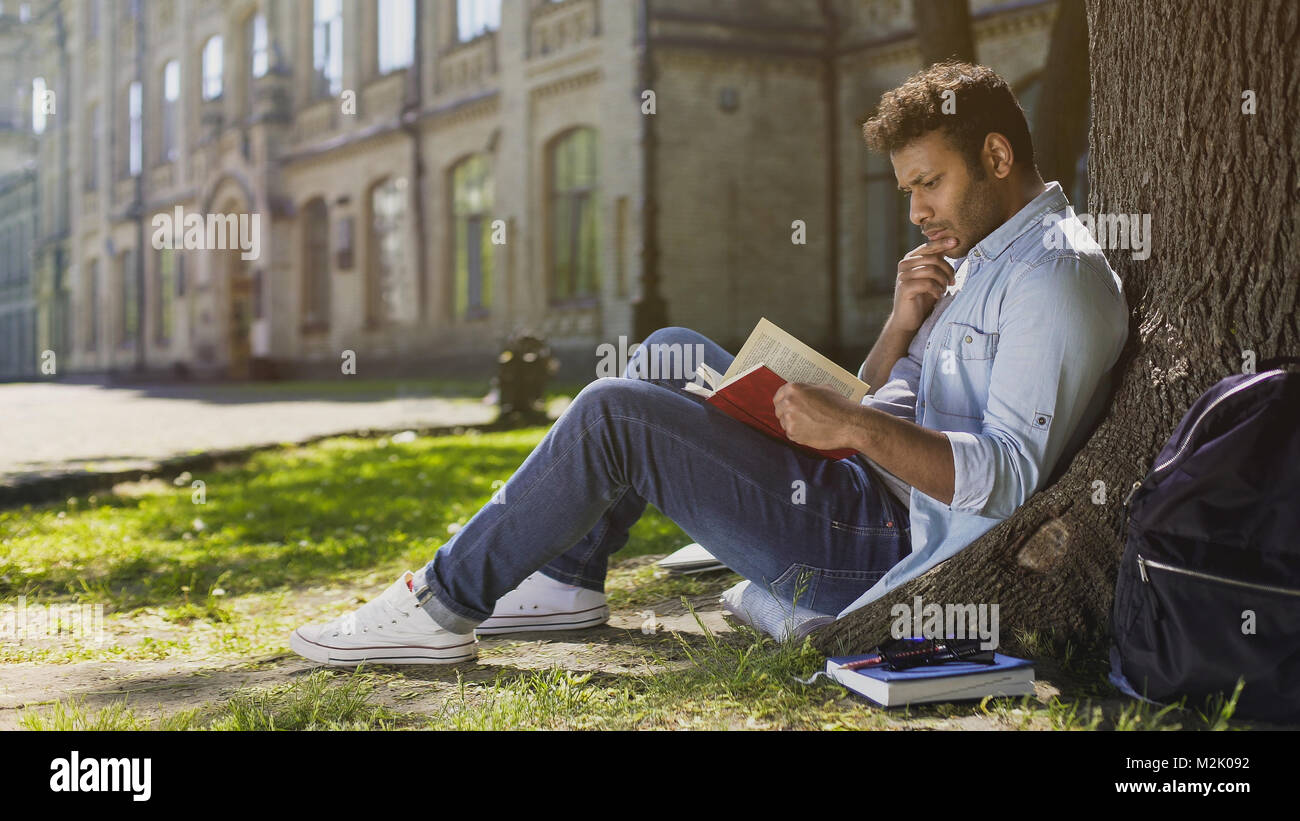 University Student sitting under tree reading book avec intrigue captivante, absorbé Banque D'Images