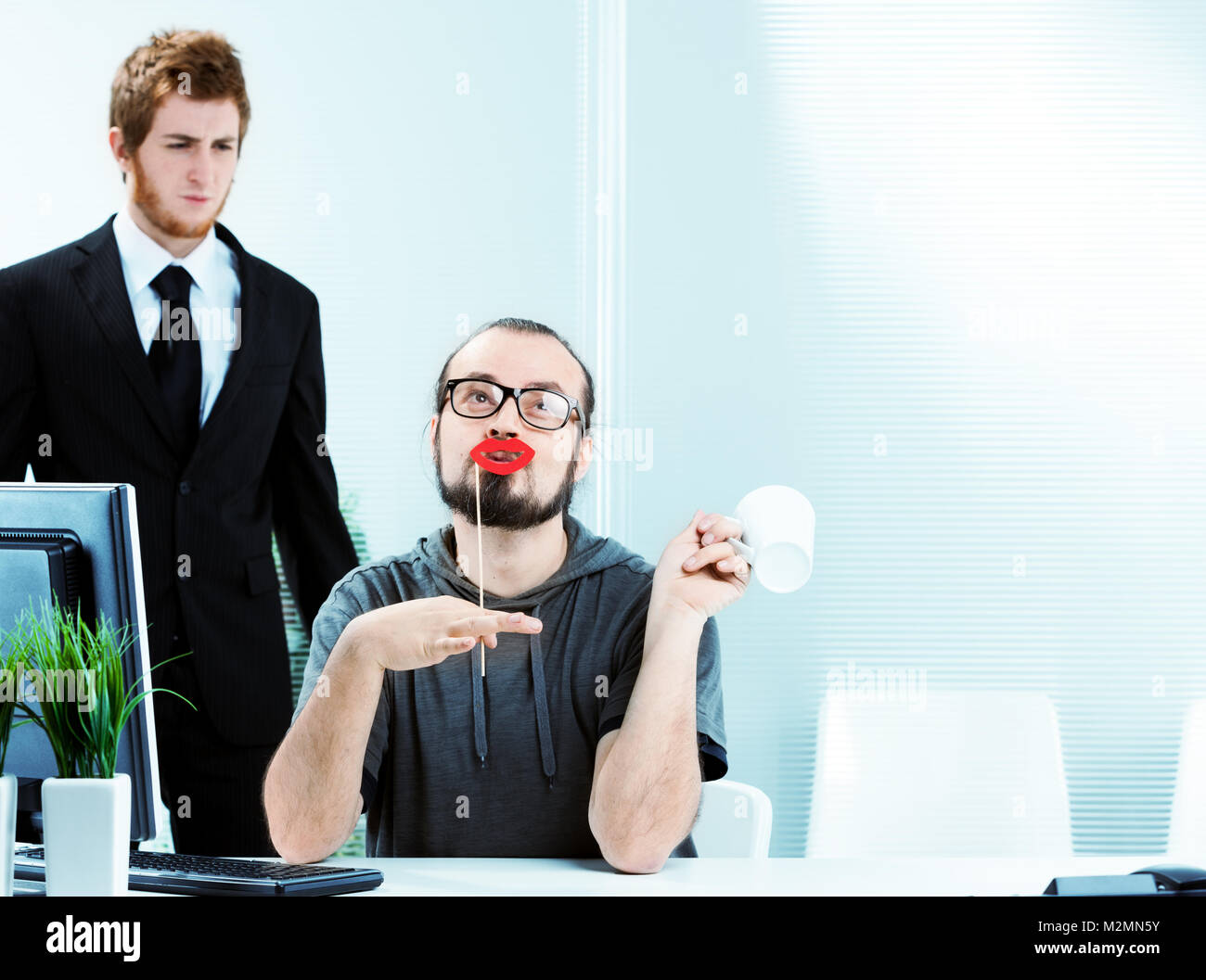 Un homme efféminé office worker wearing rouge à lèvres rouge vif, assis à son bureau simpering regardé avec dégoût par un collègue dans un élégant costume et cravate Banque D'Images