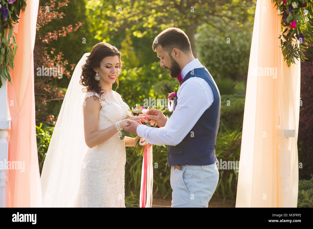 Groom bague d'usure dans le doigt de son épouse unter l'arche avec textile couleur pêche Banque D'Images