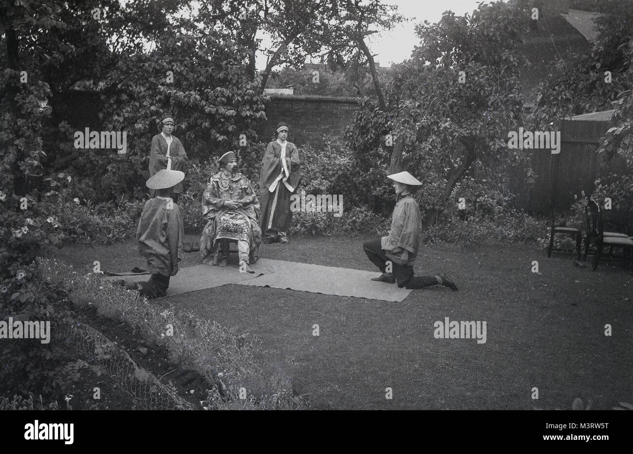 Années 1930, historique, un petit groupe de jeunes gens habillés en costumes orientaux ou chinois et portant des chapeaux de soleil ou du riz, de l'exécution d'un jouer dans un jardin de banlieue, England, UK. Banque D'Images