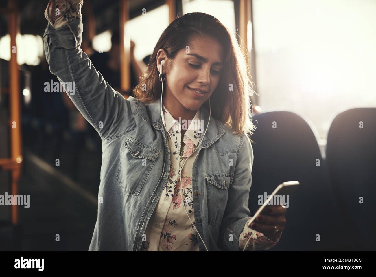 Smiling young woman wearing earphones debout par elle-même sur un bus d'écouter de la musique sur un smartphone Banque D'Images