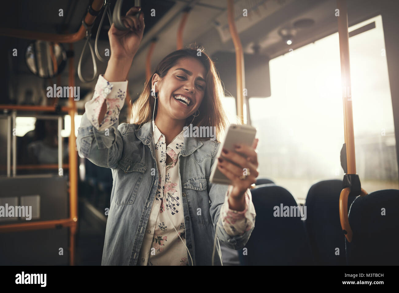 Young woman wearing earphones rire à un message texte sur son portable tout en montant sur un bus Banque D'Images