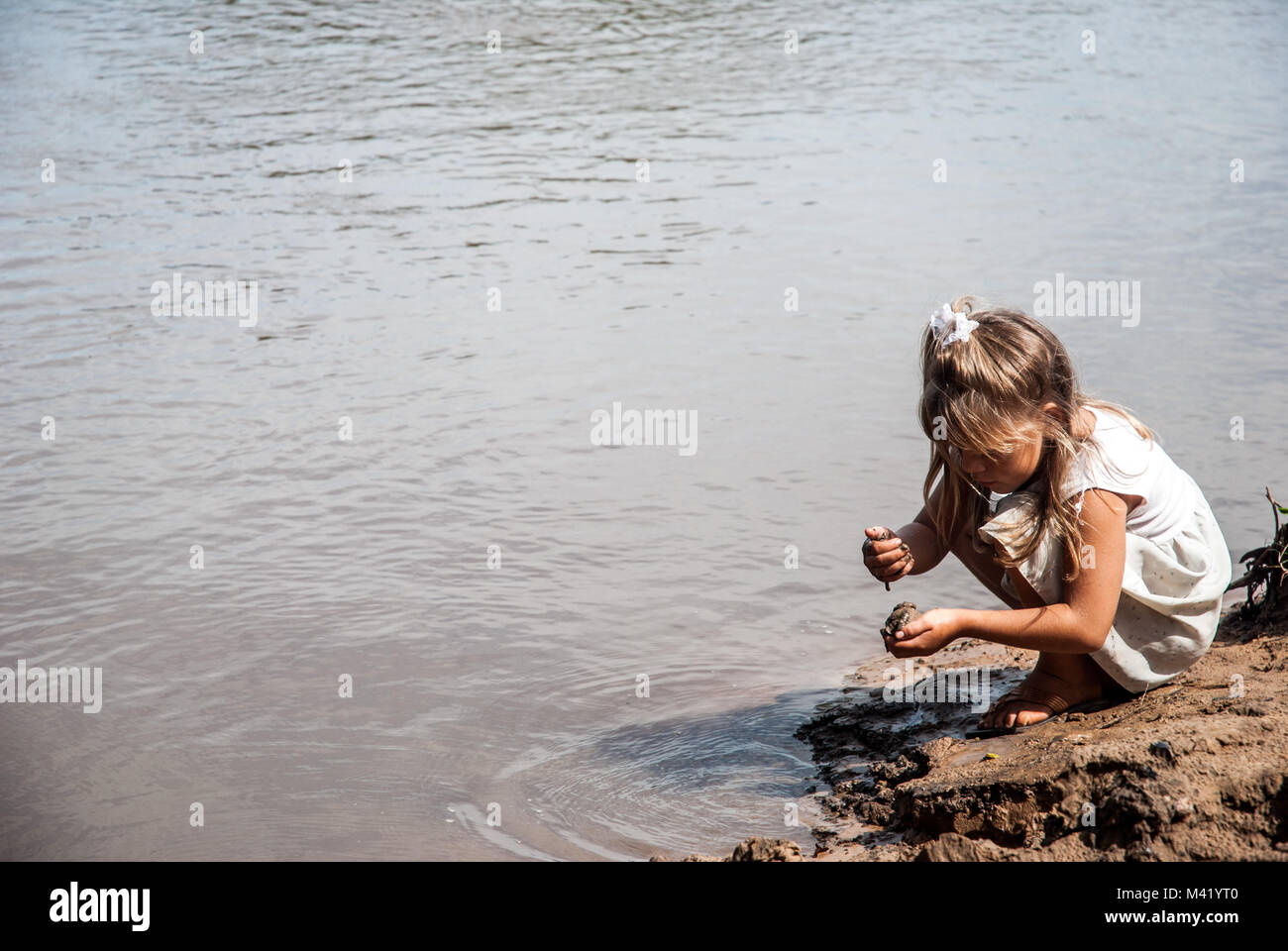 Une jeune fille blonde accroupi sur une berge et jouer avec la boue Banque D'Images