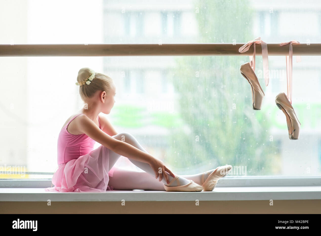 Ballerine en robe rose assis sur la fenêtre-sill. Belle jeune fille de ballet à la à la fenêtre. Paire de pointes accroché sur barre de ballet. Banque D'Images