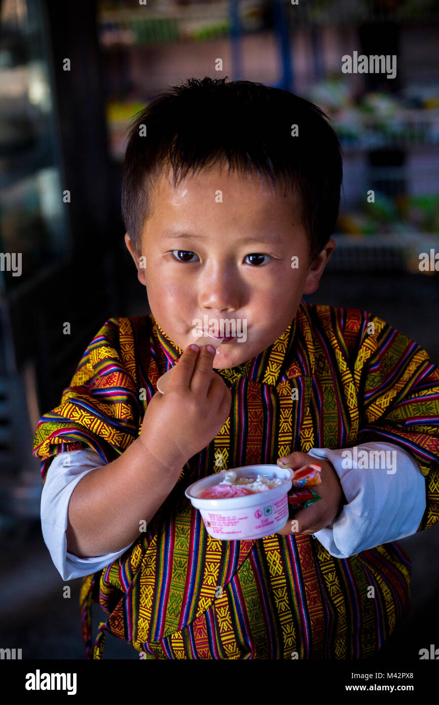 Punakha, Bhoutan. Jeune garçon en vêtement masculin traditionnel Gho, manger de la crème glacée dans le marché Lobeysa. Banque D'Images
