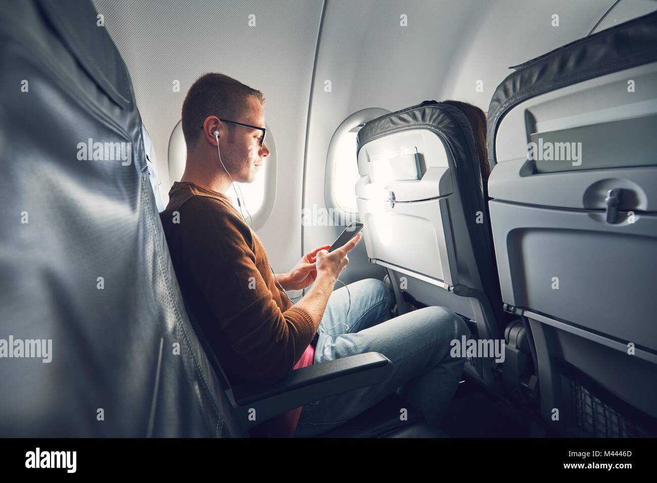 Connexion dans l'avion. Jeune homme (meilleur) à l'aide de smart phone pendant le vol et l'écoute de la musique. Banque D'Images