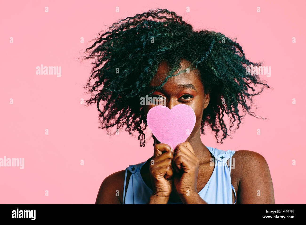 Fille aux cheveux noirs frisés bleuté tenant un cœur rose en face de son visage Banque D'Images