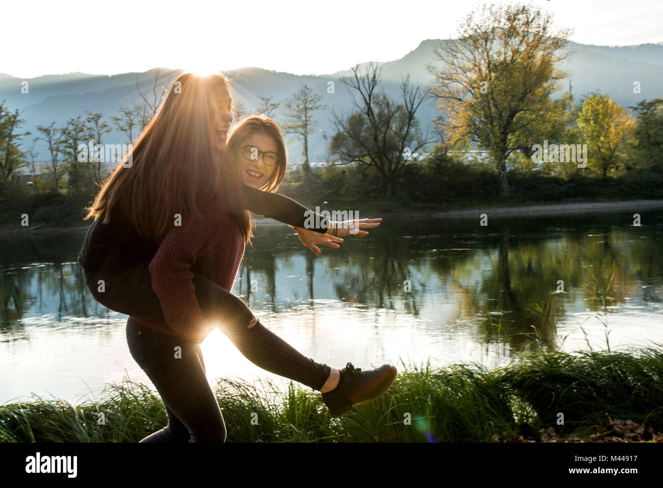 Jeune femme donnant meilleur ami piggy back on riverbank, Calolziocorte, Lombardie, Italie Banque D'Images