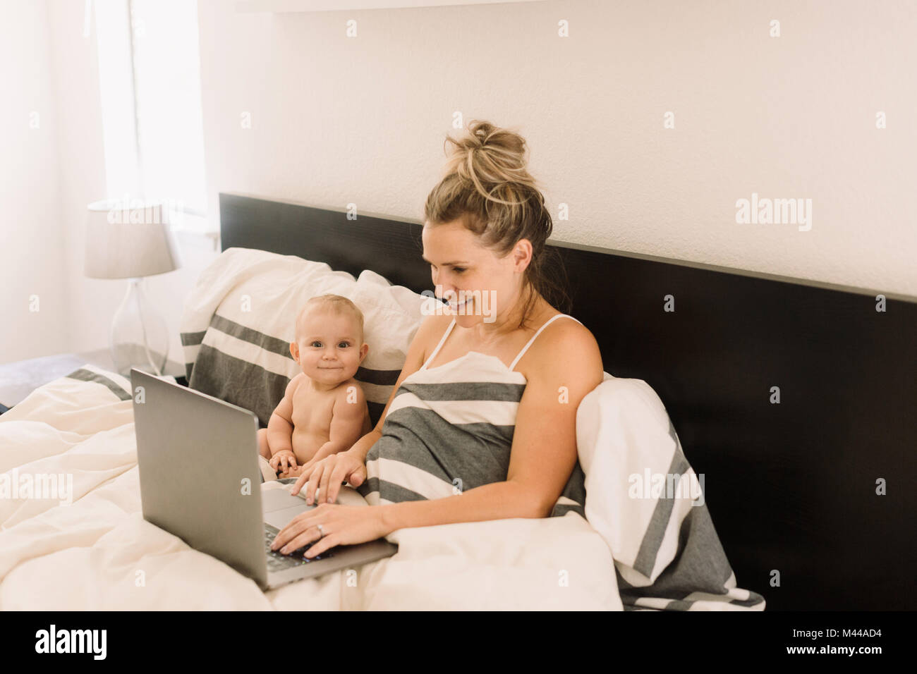 Portrait of cute baby girl sitting jusqu'au lit alors que mère using laptop Banque D'Images