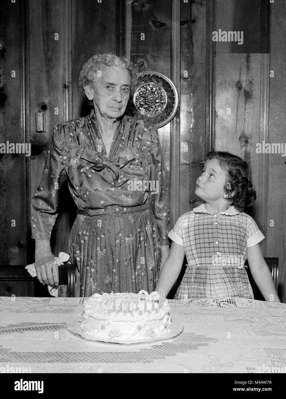 Quatre-vingt-dix ans, femme apparaît appréhension à un gâteau d'anniversaire bien que considérés par son arrière-petite-fille, ca. 1960. Banque D'Images