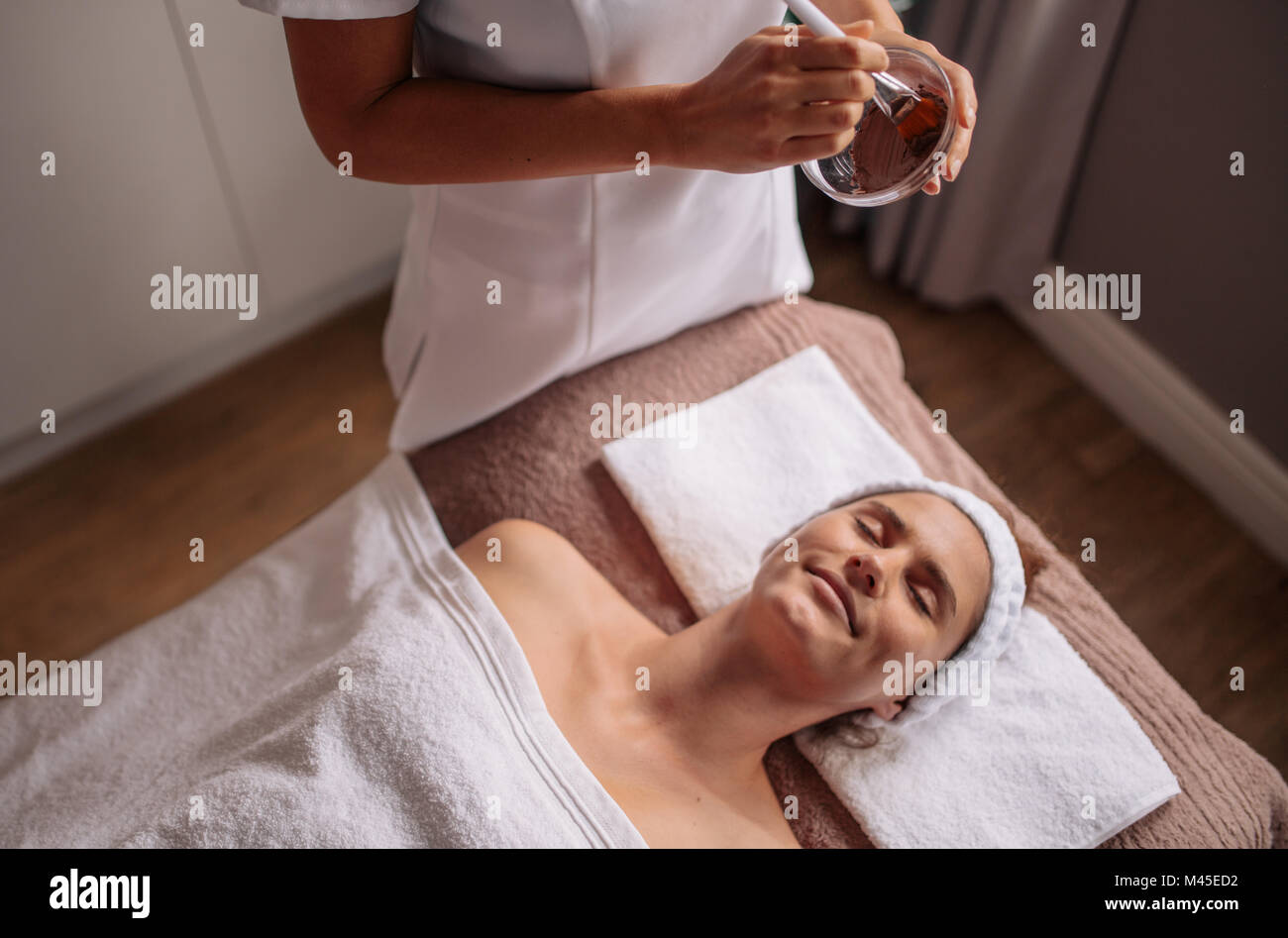 Woman lying on massage table esthéticienne avec la préparation d'un masque de visage dans un bol. Les femmes d'obtenir un traitement facial mask at beauty spa. Banque D'Images