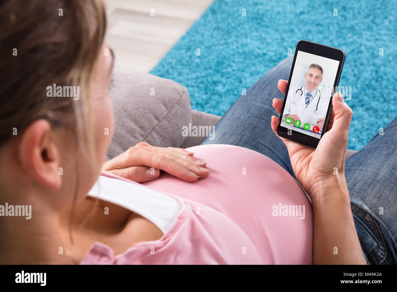 S'attendant pregnant woman Sitting on Sofa la visioconférence avec médecin sur téléphone mobile Banque D'Images