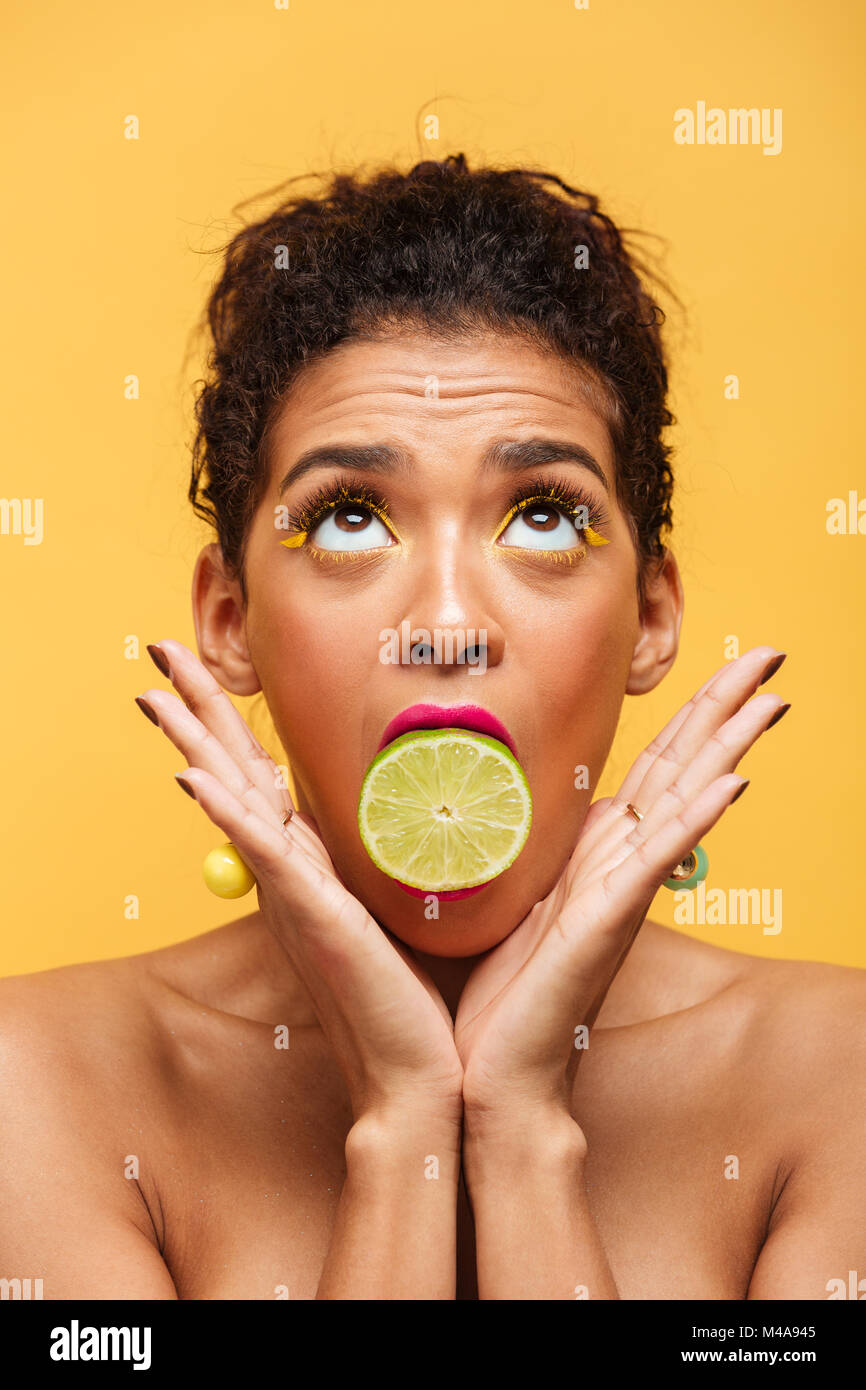 Vertical image de femme mignon avec maquillage à la mode maintenant la moitié du citron vert frais en bouche plus isolé sur fond jaune Banque D'Images