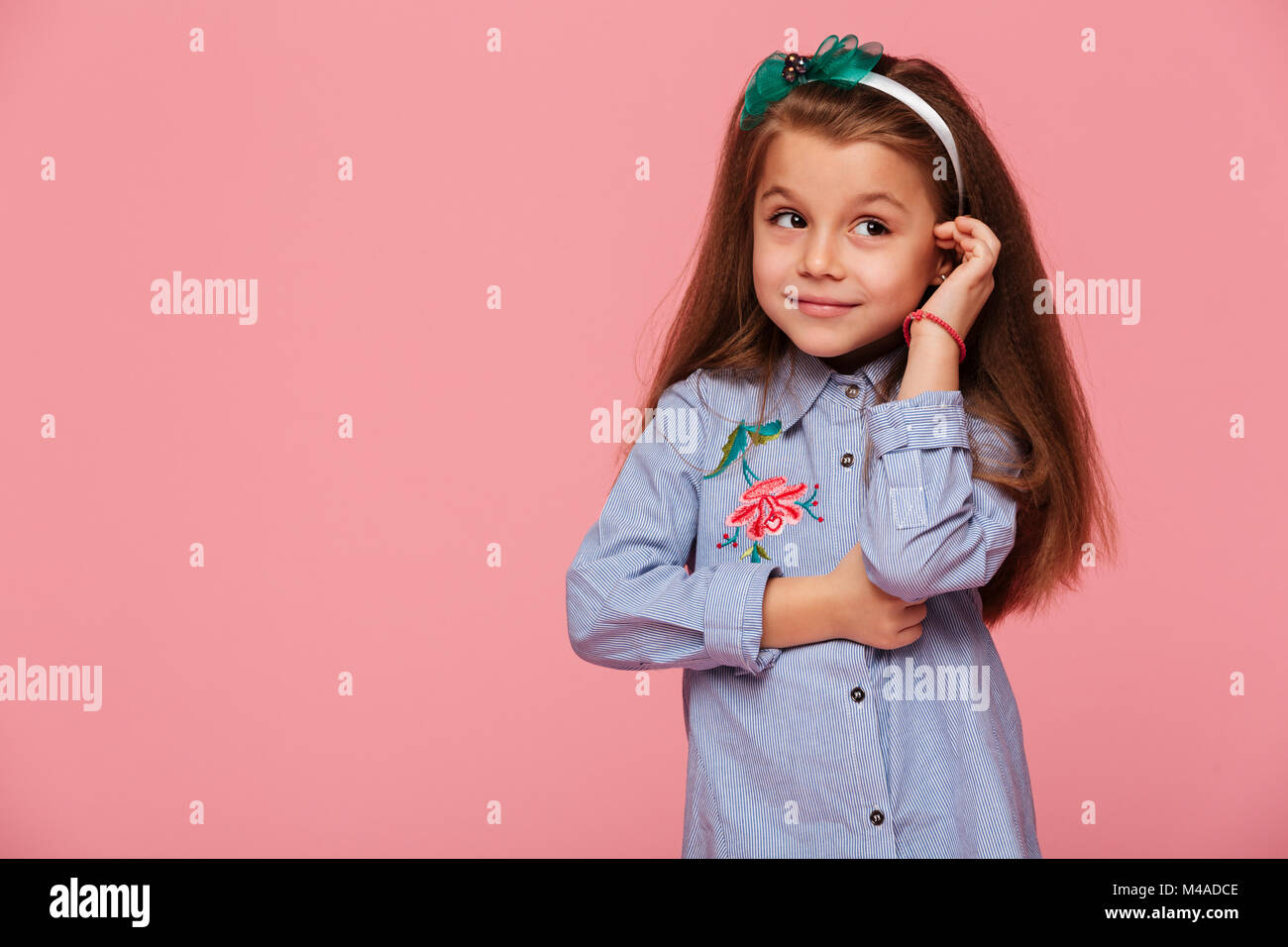 Portrait of cute schoolgirl 5-6 ans toucher ses longs cheveux auburn avec genre beau sourire, isolé sur fond rose Banque D'Images