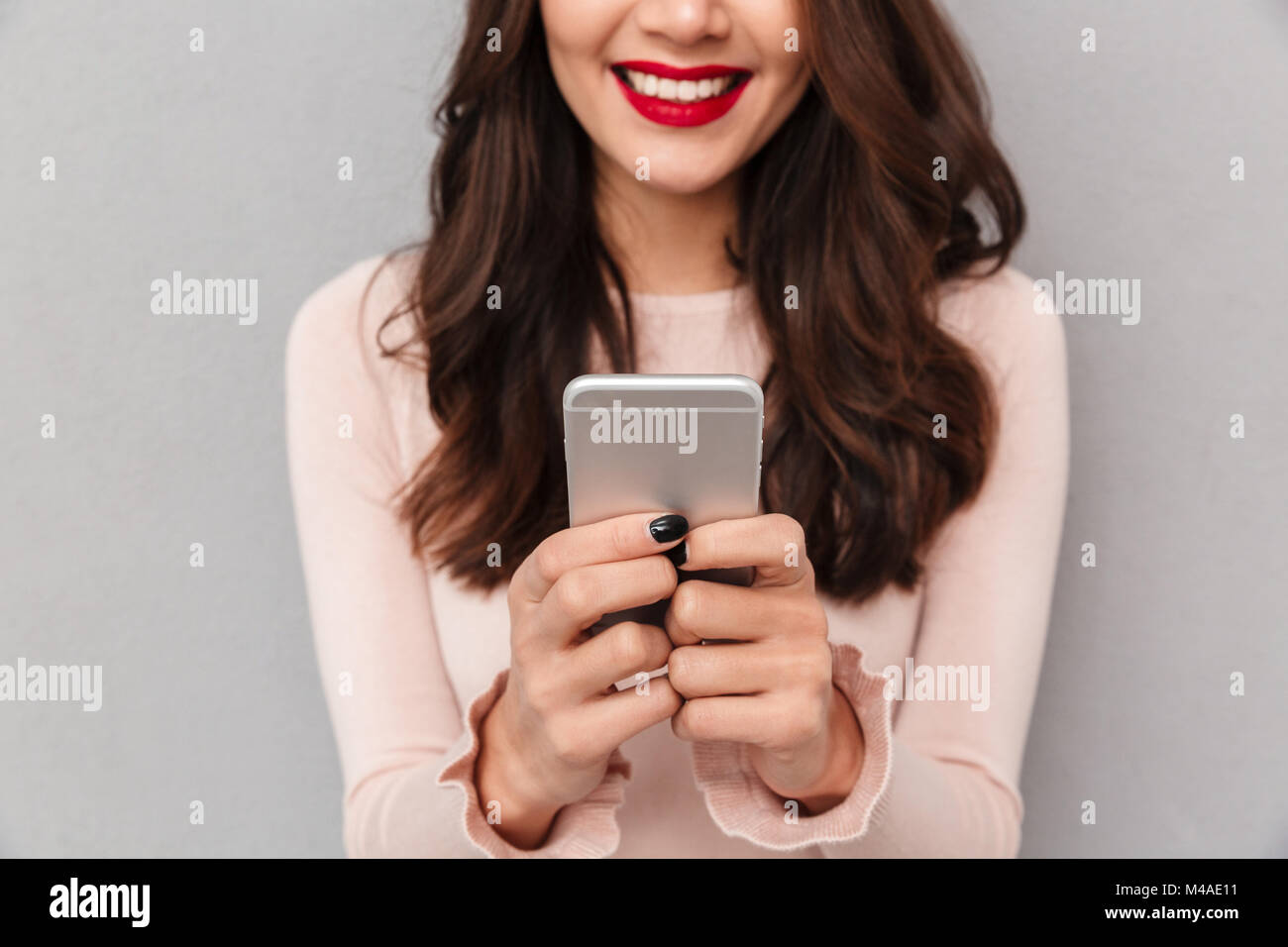 Close up image of smiling female with red lips holding mobile phone chatter ou jouer à des jeux via appareil moderne, sur fond gris, cropped Banque D'Images