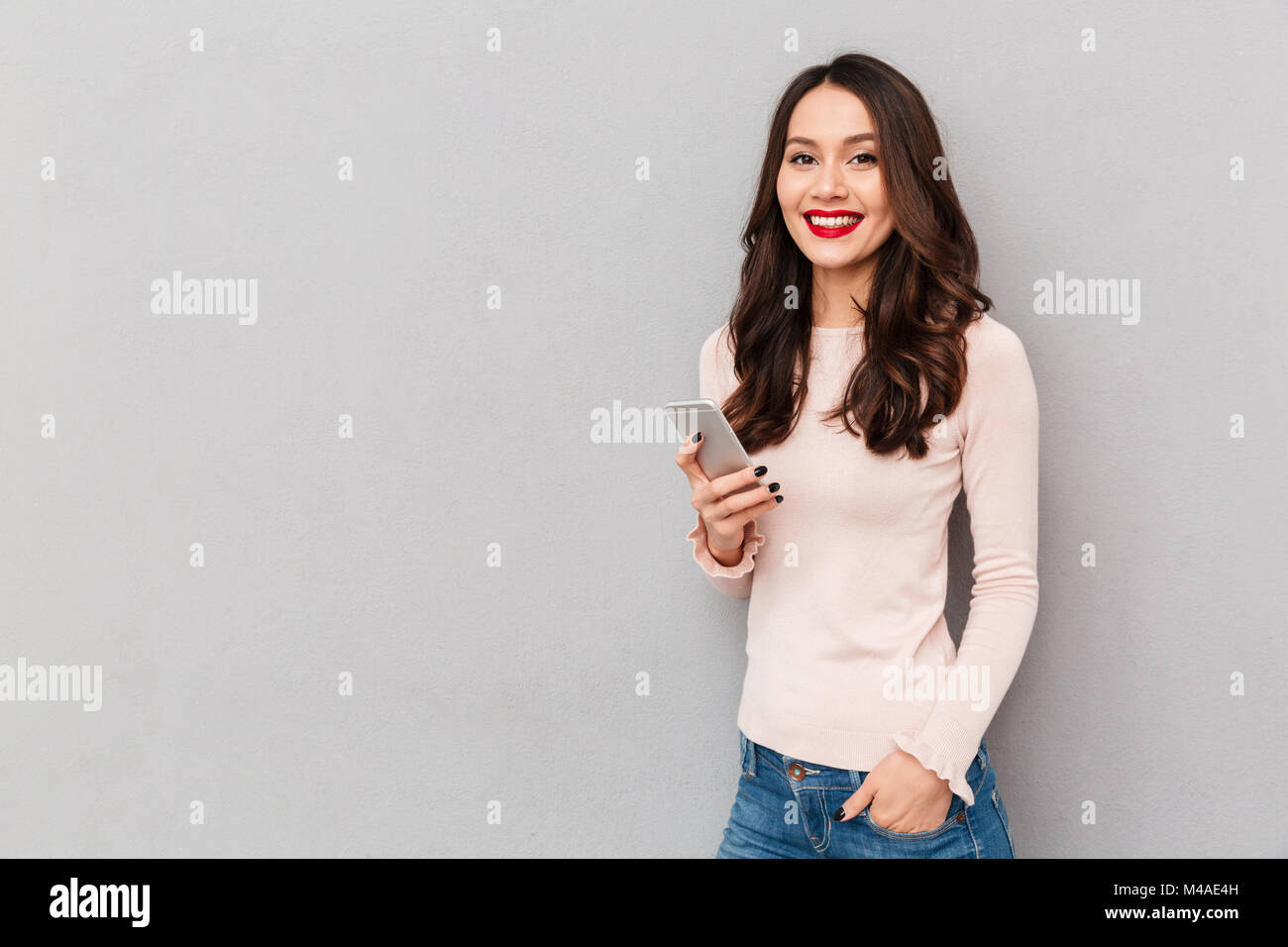 Photo horizontale de smiling woman with red lips saisissez du texte ou message défilement dans réseau social, à l'aide de blackberry sur fond gris Banque D'Images